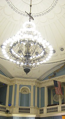 A room with high ornate blue-and-white walls and a white-domed ceiling, from which a large chandelier hangs by a rod. Along the rod, between the chandalier and the ceiling, is the figure of a fish.