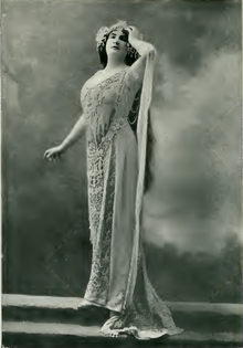 A white woman, standing, hand on forehead, wearing a gown and headdress typical of 1900s Wagnerian opera.