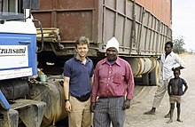 Ted Conover stands with a Kenyan man next to a large industrial truck parked on a dirt road while a younger Kenyan man and little boy look on.