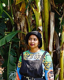 Portrait of Youlendree Appasamy standing in front of banana trees. Youlendree is a feminist free radical, a writer, collagist and zine-maker who makes work about and with Indian indentured communities in South Africa.