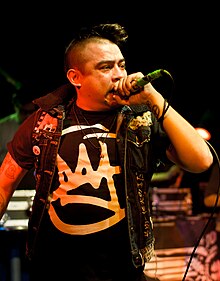 A man with a mohawk in a black T-shirt and open black jacket holds a microphone close to his mouth