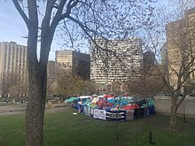 A dense group of tents on a university lawn in downtown Montreal, surrounded by fences bearing banners