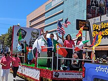 A parade float going down a street. There is a large soccer ball. Station employees are on the float waving flags of various Spanish- and Portuguese-speaking countries, and there is promotion for the 2022 FIFA World Cup.