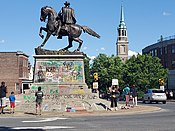 An equestrian statue covered in spray-painted graffiti with a protester in front holding a sign reading "BLACK LIVES MATTER".