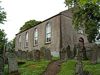 A plain chapel-like building with Georgian-style windows on a hill, surrounded by a dense churchyard.