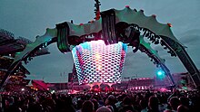A concert stage; four large legs curve up above the stage and hold a video screen which is extended down to the band. The legs were lit up in green. The video screen has multi-coloured lights flashing on it. The audience surrounds the stage on all sides.