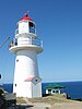 White conical lighthouse with red dome, a green roofed structure seen at the far background