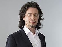 Picture of a man in his mid-forties with long, tousled hair wearing a jacket and a shirt with an open collar