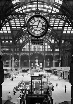 Large high-ceilinged space with round iron arches, covered in glass-dome skylights, floored with vault lights, clock central in foreground.