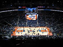 An arena packed with spectators. A basketball court is in the center, and a scoreboard is directly above it.