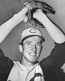A man in a white baseball jersey with dark sleeves and a white cap with a dark "C" on the front holds his hands above his head in a glove.