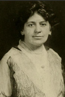 A white woman with dark curly hair, wearing a lace-trimmed dress or blouse