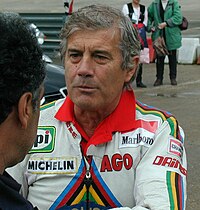 A man wearing a white leather top with writing on it. His arms are crossed and there is a man standing in front to his left.