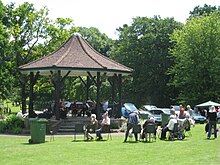 Grassy area with people sitting in chairs in front of a bandstand with tiled roof.