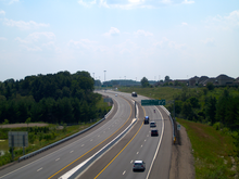 A four-lane divided highway (dual carriageway) curves over a wooded stream. A subdivision looms on a hill in the distance.
