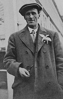Picture of Herbert Whitley facing towards the camera, holding a tobacco pipe in his right hand down near his waist, and with an exotic flower in his left lapel
