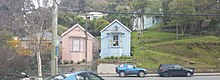 Two almost identical small wooden houses next to each. A pink one and a blue grey one. Set up from the street with a concrete retaining wall. A steep grassy bank is to the right. Parked cars and street posts in the foreground.