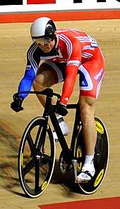 Chris Hoy wearing a bicycle helmet, visor, cycling shorts and top cycling on a racing bike in a velodrome.