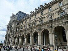 A classical building with ornamental design rises above a small crowd. Rounded archways line the front of the structure.