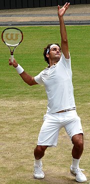 A brown-haired man in a white polo shirt raises his left arm, preparing to serve