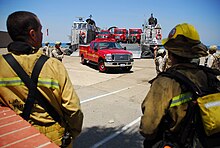 Fire Department vehicle driving off landing craft with firefighters standing in foreground