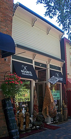 An off-white wooden building attached to a brick building on the left and a slightly similar building on the right, seen at an angle to the right. At street level is a storefront for "Noori's Collection" with carpets and statues displayed in front. Above it is a plain wooden front between two bracketed wooden projections.