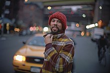 A man with facial hair, dressed in a flannel jacket and knitted hat, and a city street in the blurred background