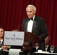 "An elderly man in formal wear makes a speech at a podium during a dinner party."