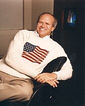 Dick Ebersol, wearing a white shirt with a USA flag on it, smiling widely, sitting in a black chair.