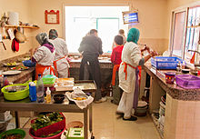 3 trainees dressed in white, cooking with the restaurant's chef who is dressed in black. A young volunteer is taking the food to serve to customers.