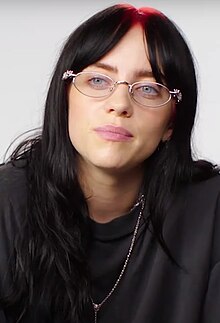 Artist has pale eyes and a smile, jet black hair, ornate wireframe glasses, necklaces, and a punky black top
