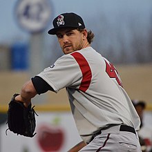 A man wearing a gray baseball jersey with a red stripe under the arm and red numbers on the back and a black cap and glove prepares to pitch a ball.