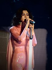 A young brunette woman singing into a microphone while playing keyboards. She is wearing a pink unitard and has a headpiece with a pair of purple Minnie Mouse-like ears.