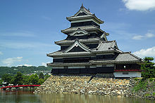 A large 5-storied castle tower with black wooden walls located on a platform of unhewn stones surrounded on two sides by water. The tower is connected to lower structure.