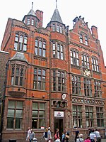 The front of a building with three storeys plus attics is seen from a slight angle. The ground floor is in stone; the upper storeys are in brick with stone dressings. On the front is a frieze containing heraldic shields, a coat of arms, and a variety of windows; at the top are two turrets and a shaped gable.