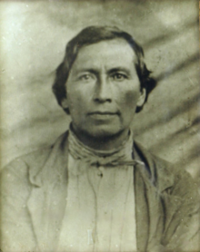 A portrait of an adult man with short, dark hair wearing a single-breasted jacket and a neckerchief.