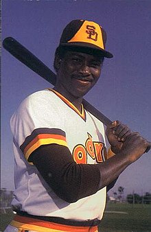 A man in a white baseball uniform and brown and yellow cap