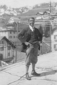 A black and white photo of a man standing on a street. He has his hands on his hips and is smiling towards the camera.
