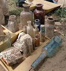 A long-necked greenish-blue bottle covered with patches of dirt lies on its side at the lip of a yellow dustpan, on which are placed upright bottles of varying size and two fragments of plaster, all also covered with dirt. Behind the dustpan stand several larger bottles, one deep red. In the background is another yellow dustpan, empty, behind which is a blue tarp atop which is a pile of the excavated dirt
