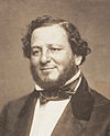 Photographic portrait of a middle-aged, curly-haired man with a short beard and a slight smile. He is formally dressed in the fashions of the mid-19th century.