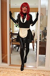 A man standing in a doorway, wearing a latex maid uniform and make-up.