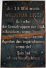 Damaged metal plaque reading "Am 3.9.1956 wurde WALDEMAR ESTEL Getreiter der Grenztruppen der NVA in Ausübung seine Dienstes an der Staatsgrenze von Agenten des Imperialismus ermordet. Sein Tod ist uns Verpflichtung."