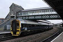 A two coach train beneath a lattice bridge