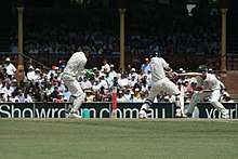 A cricket match being held. The batsman hits the ball and the other players try to catch it. The green field and the audience are visible in the far.