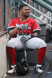 A man wearing a red jersey, gray pants, and high red socks sitting on a chair in a dugout smiling with bat in hands