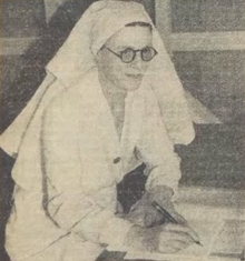 A white woman wearing glasses and a nurse's uniform sits at a desk, pen and paper in hand.