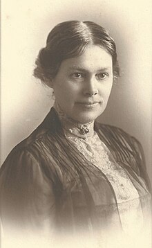 A photograph of Margaret Eliza Maltby circa 1908. She has dark hair parted down the middle and pulled back in a bun. a white lacy blouse is buttoned all the way up to her throat and secured with a brooch. a dark semi-transparent jacket overlays the blouse.