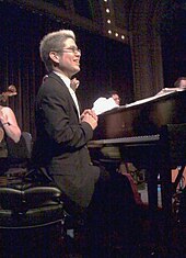 A man sitting on a piano bench behind a pian; he is wearing a suit and glasses, and his hands are folded.