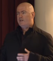 A close-up image of bald man looking up behind the camera while talking at a conference.