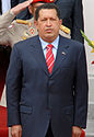 A photograph of a standing man with black hair wearing a dark dress suit, a light blue dress shirt, and a red-and-blue striped tie